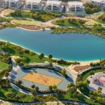 Aerial view of sports facilities including football pitch, basketball court, and volleyball court next to a scenic lake at Violet 3 in DAMAC Hills 2, Dubai