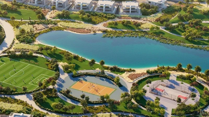 Aerial view of sports facilities including football pitch, basketball court, and volleyball court next to a scenic lake at Violet 3 in DAMAC Hills 2, Dubai