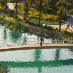Wooden pedestrian bridge over scenic water stream surrounded by lush green parks and palm trees at Violet 3 in DAMAC Hills 2, Dubai