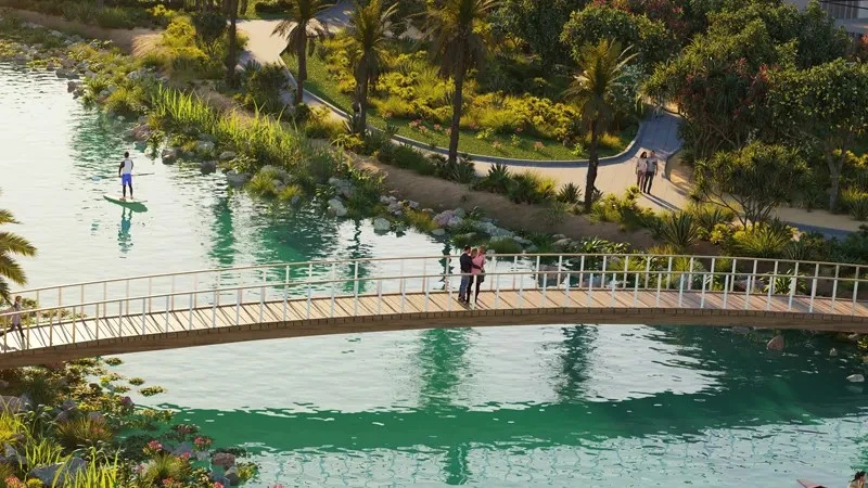 Wooden pedestrian bridge over scenic water stream surrounded by lush green parks and palm trees at Violet 3 in DAMAC Hills 2, Dubai